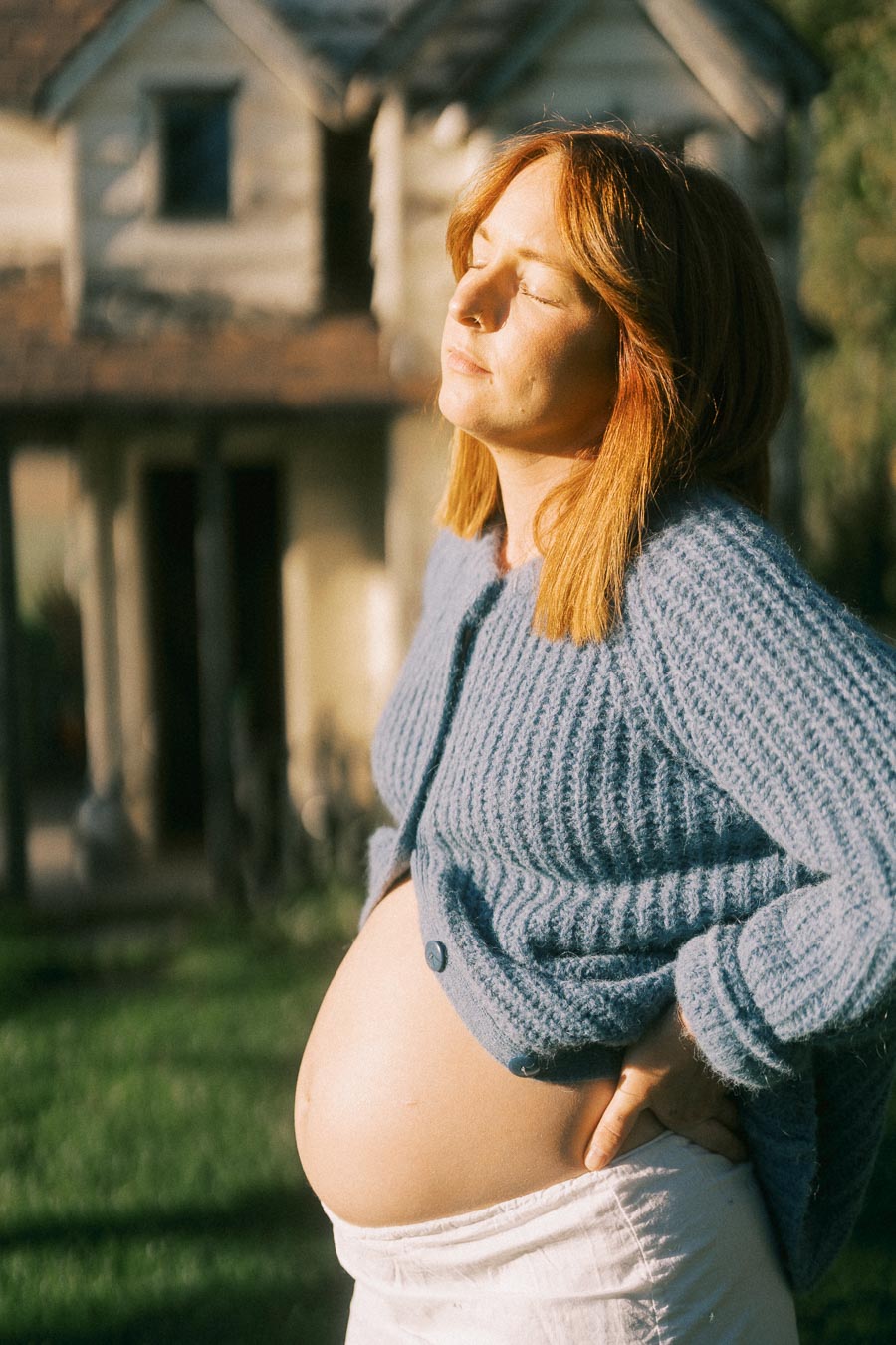 A pregnant woman standing outside a house, basking in sunlight with a peaceful expression, wearing a blue cardigan and white skirt.