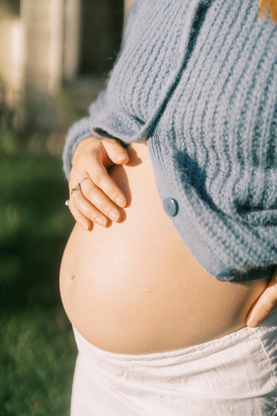 A close-up image of a pregnant belly with a woman's hand gently resting on it, wearing a cozy blue knit sweater and standing outdoors in a sunlit garden.