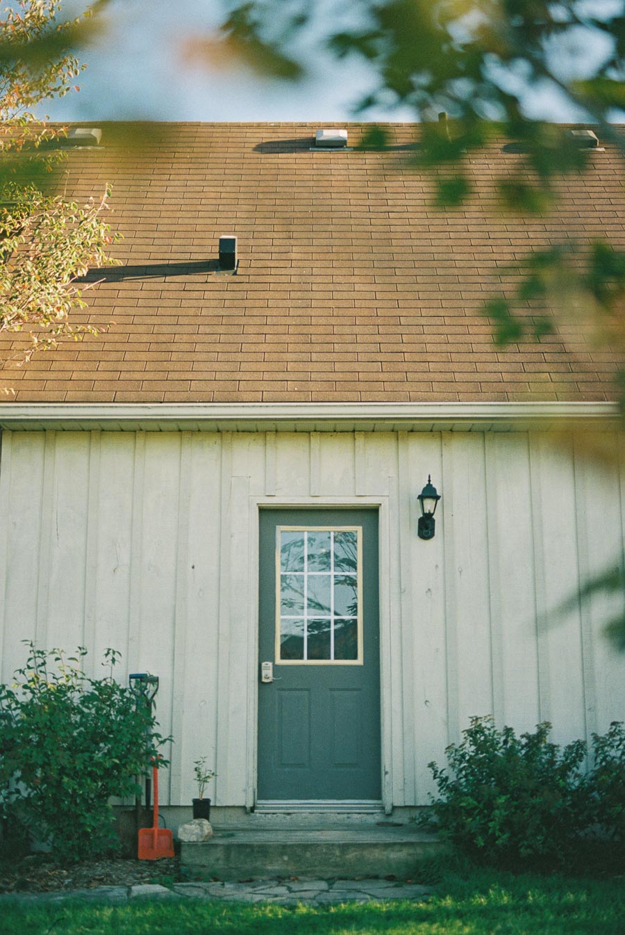 Charming wooden house exterior with a brown shingle roof and a green framed glass door, surrounded by greenery and a rustic garden shovel by the entrance.
