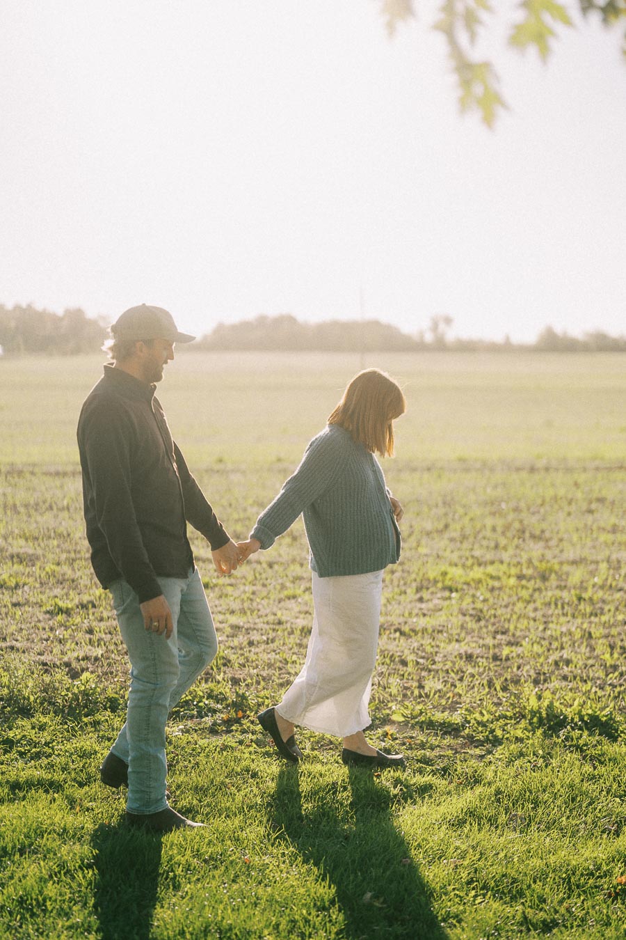 Couple walking hand in hand in a grassy field during daylight, with the sun illuminating the serene landscape. Both are wearing casual clothing, and the image conveys a peaceful and intimate outdoor moment.