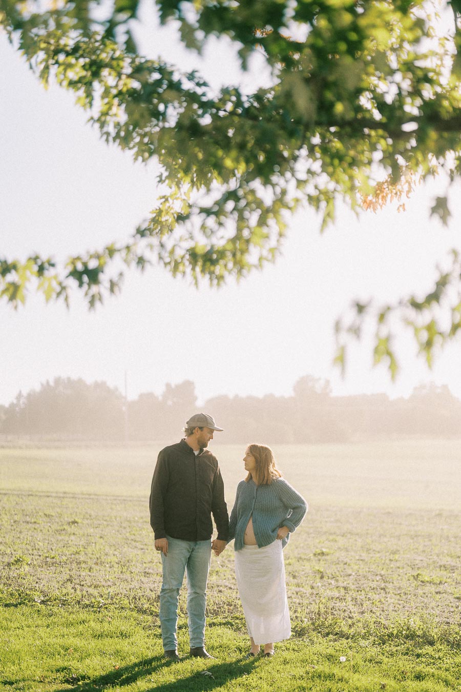 A couple holding hands in a sunlit field, with green leaves framing the image, showcasing an intimate outdoor moment.