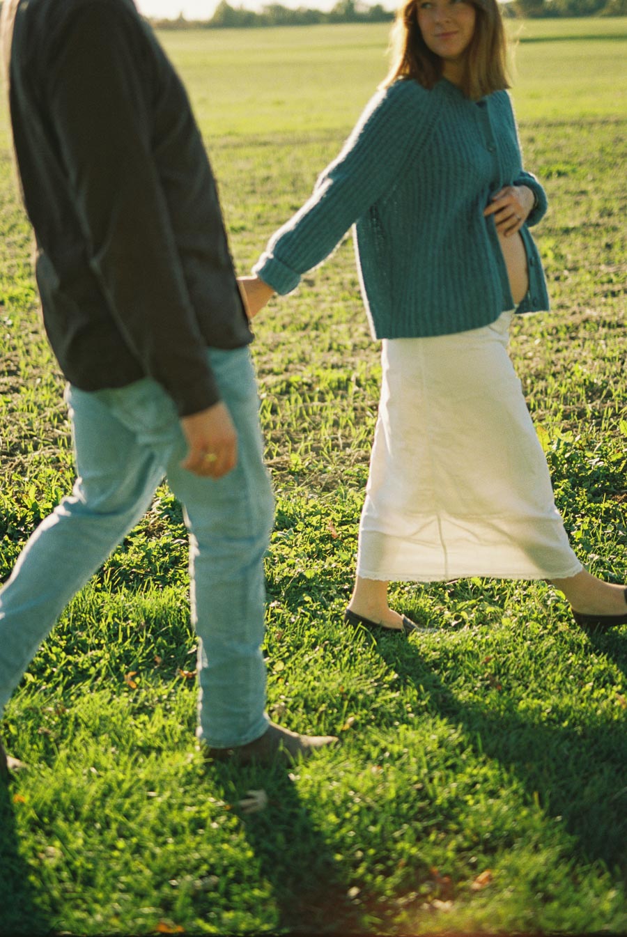 A couple holding hands while walking in a sunlit field, with the woman gently touching her pregnant belly, expressing love and anticipation.