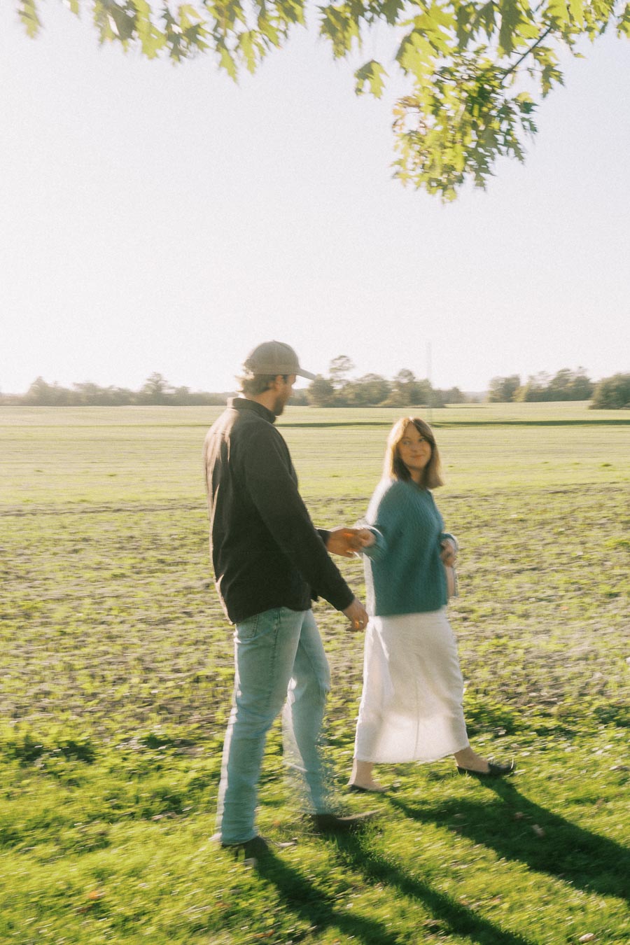 A couple strolling hand in hand through a sunny green field, surrounded by trees, enjoying a leisurely afternoon outdoors.