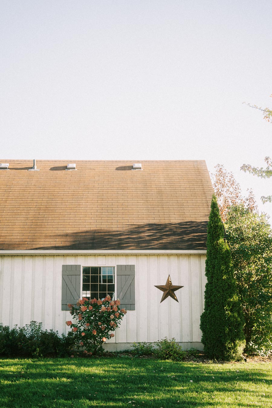 Charming countryside house with beige siding and gray shutters, featuring a decorative barn star and blooming shrubbery under a clear blue sky.