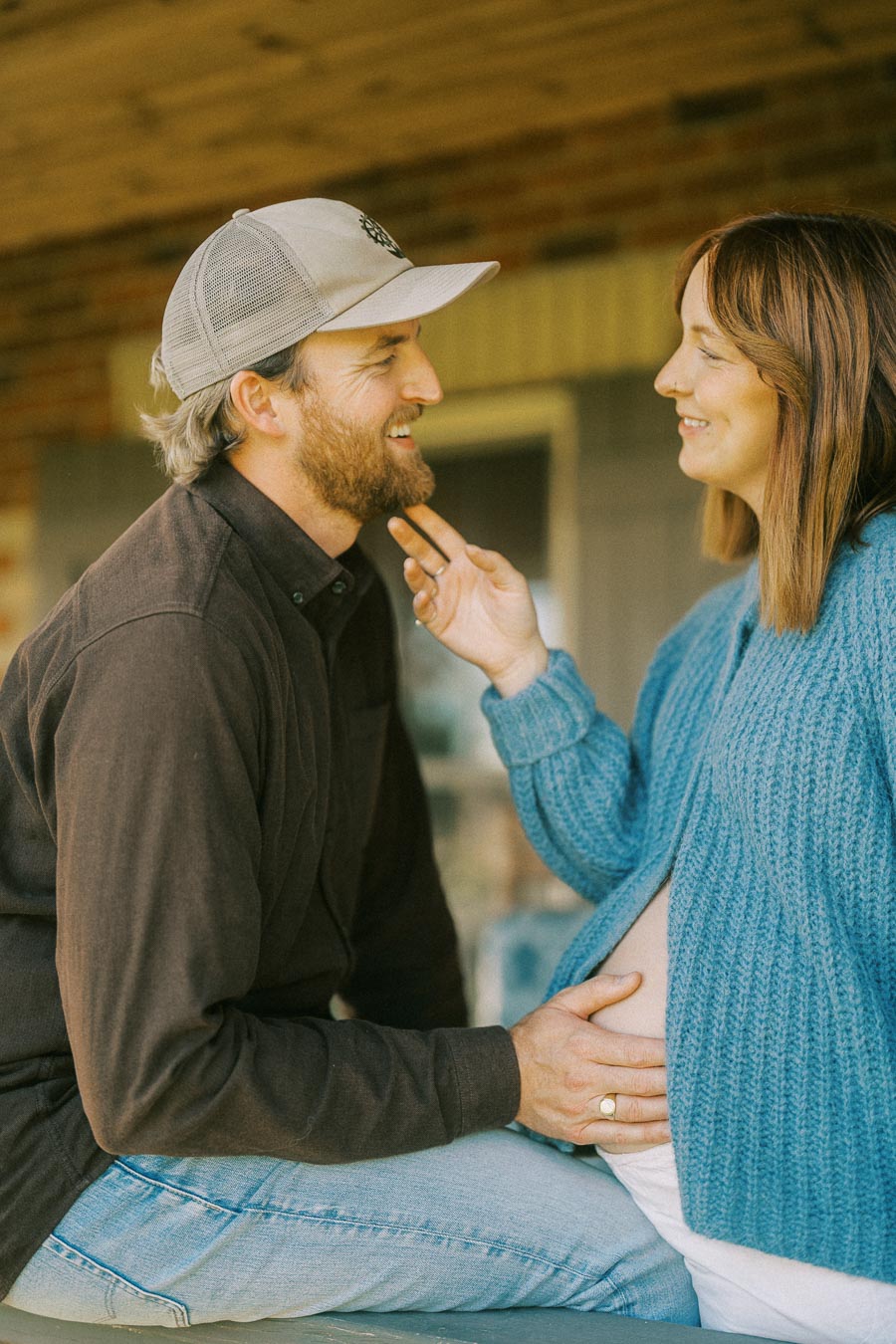 Young couple smiling and embracing as they sit outdoors, with the woman cradling her baby bump and the man wearing a casual cap.