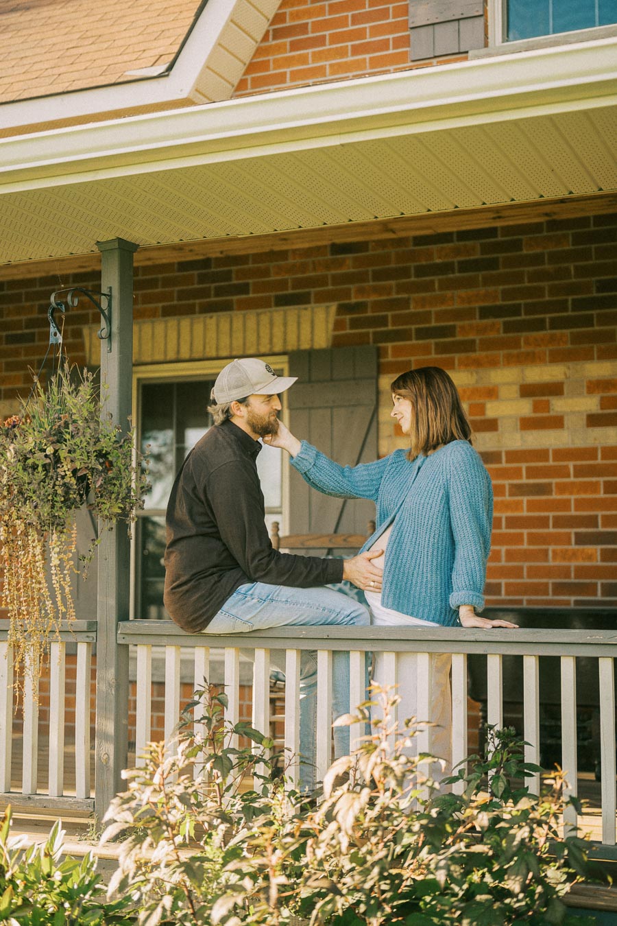 A couple sitting on a porch, with the man wearing a cap and casual clothes, gently touching the woman's face. The woman, dressed in a blue sweater and holding her pregnant belly, is smiling at him. They are surrounded by a hanging plant and garden foliage, with a brick house backdrop.