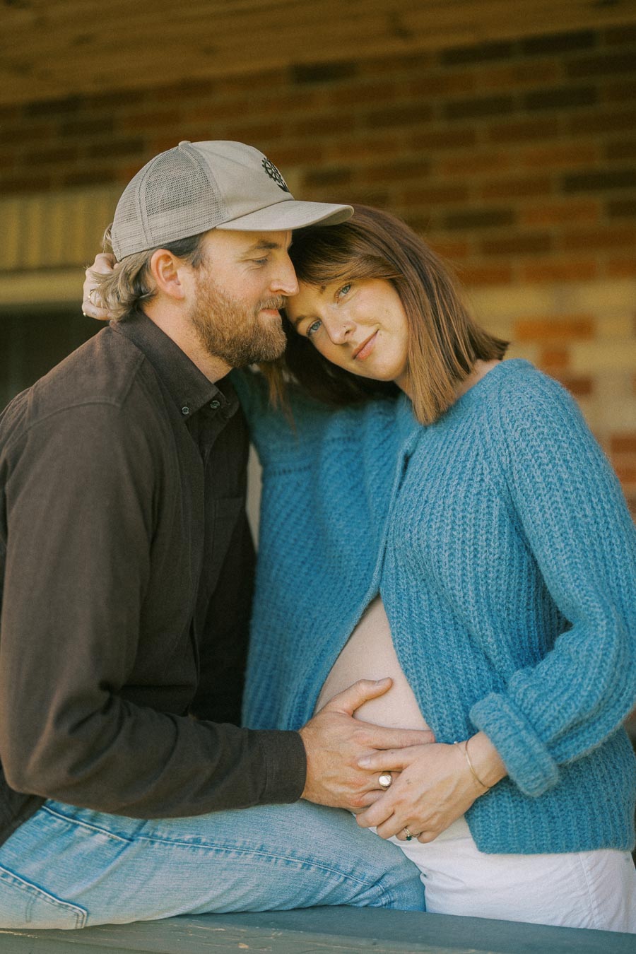 Couple embracing with woman in blue sweater showing baby bump; outdoor maternity photo with warm, intimate atmosphere.