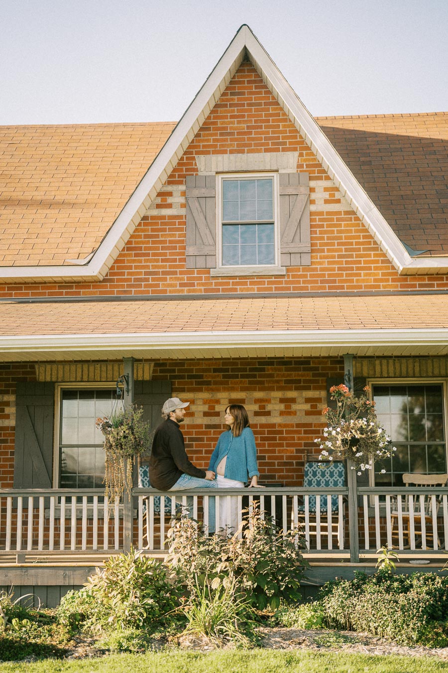 A couple engaged in conversation on the porch of a charming brick house adorned with shutters and hanging plants, basking under clear blue skies.