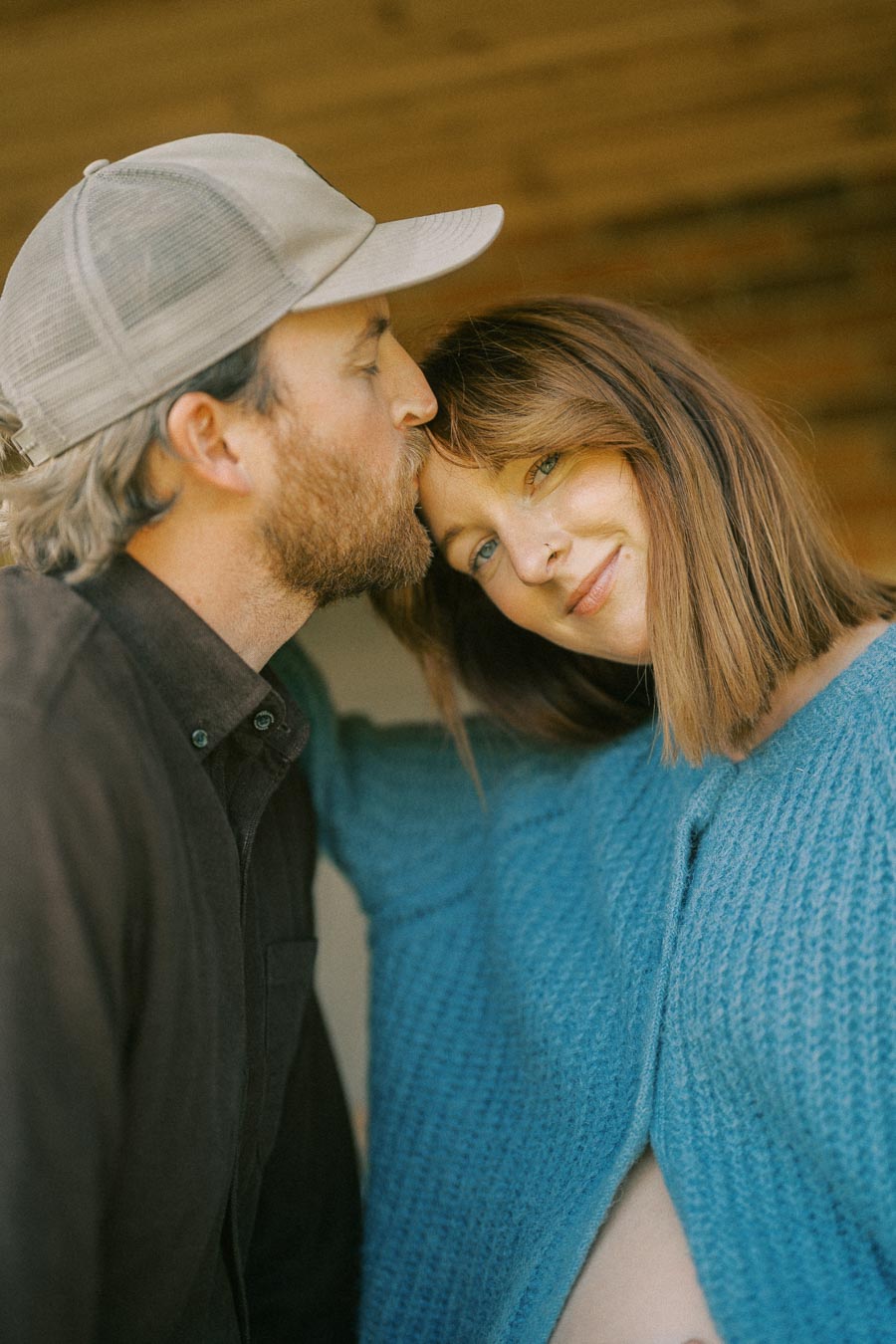 A loving couple shares a tender moment as a man in a hat kisses a smiling woman's forehead, with her wearing a blue sweater, conveying warmth and connection.