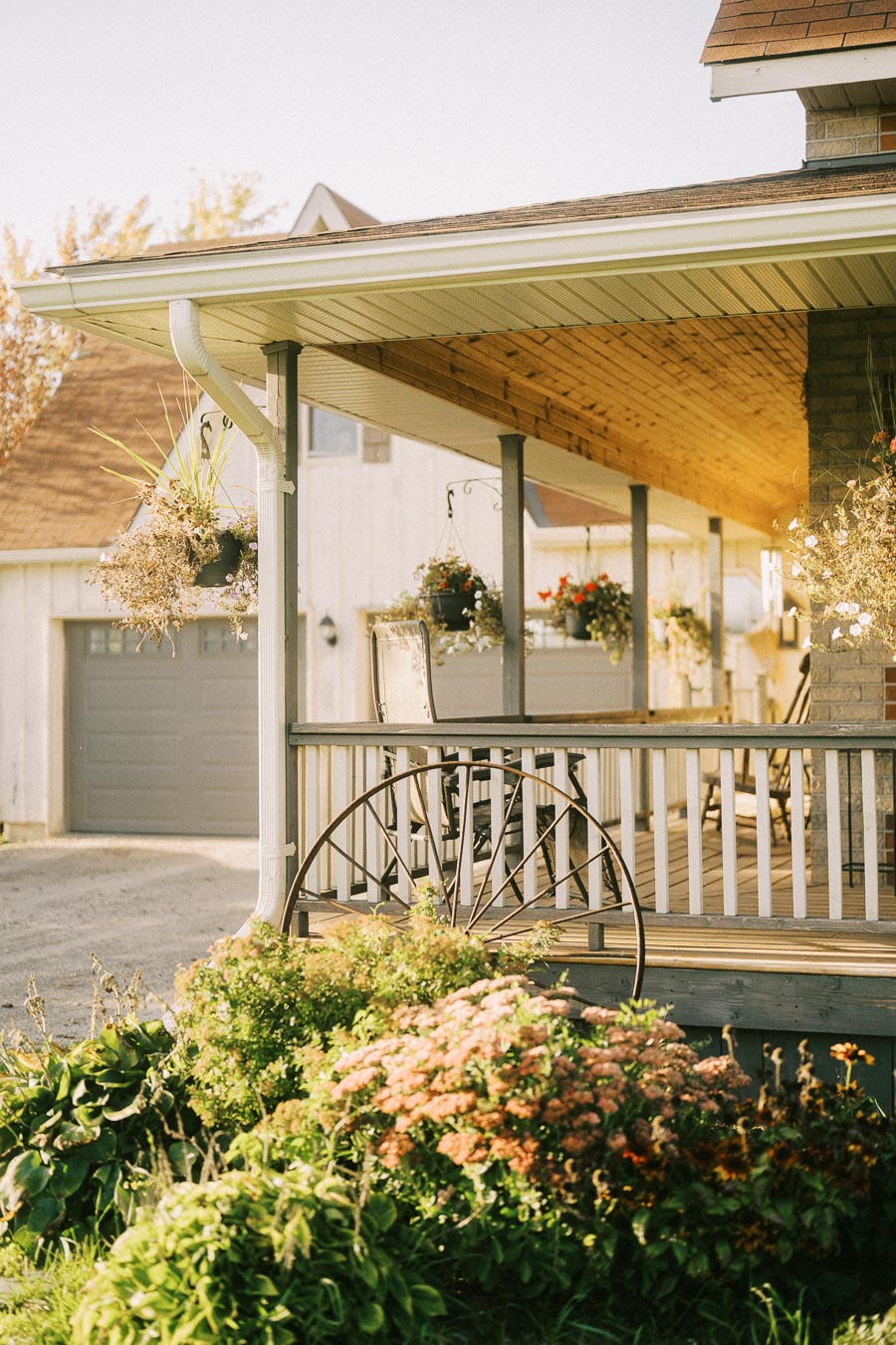 Charming rural porch with hanging flower baskets and garden plants, featuring a vintage wagon wheel, under a slanted wooden roof on a sunny day.