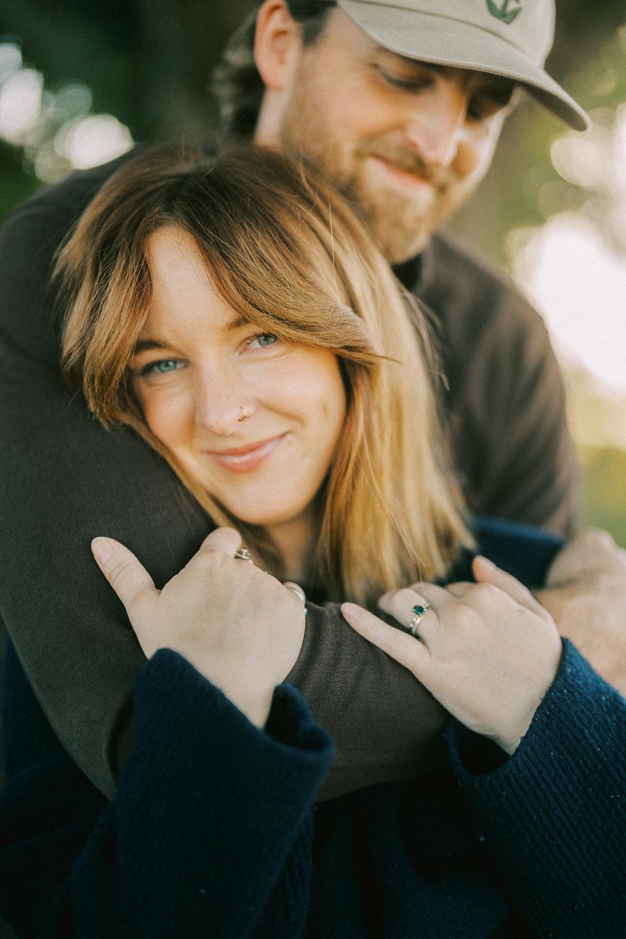 A smiling woman with long hair is embraced by a man in a casual outdoor setting, depicting warmth and affection.
