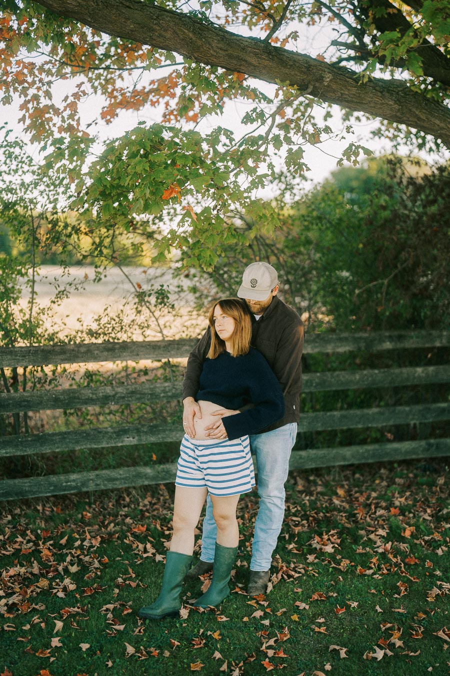 Young couple embracing under a tree in a serene outdoor setting, with autumn leaves scattered on the grass, showcasing tenderness and togetherness.