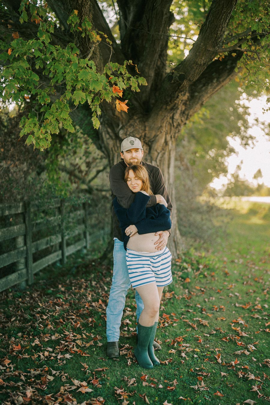 Man embracing a pregnant woman in striped shorts while standing under a tree, surrounded by autumn leaves.