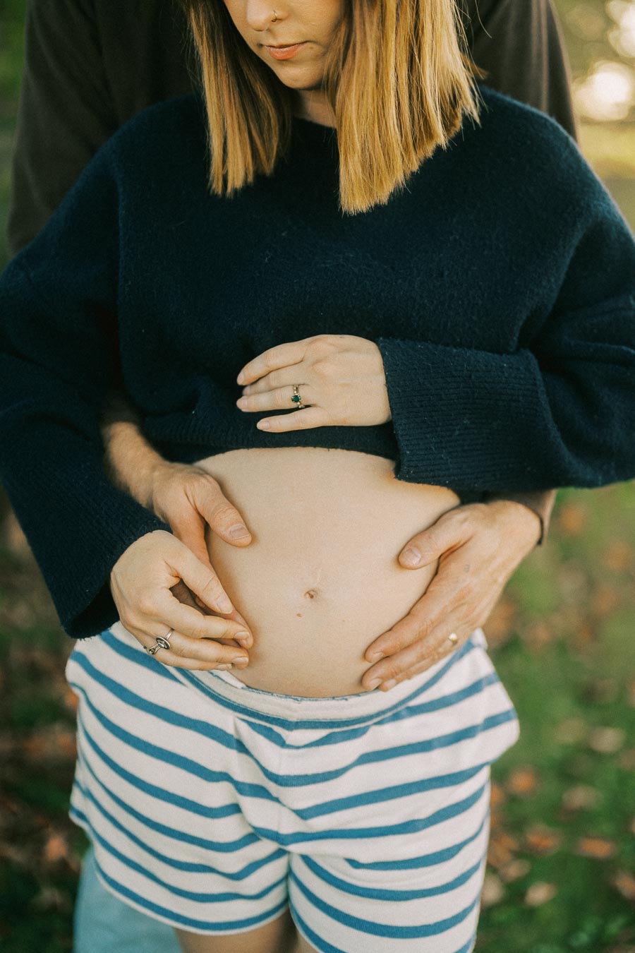 A pregnant woman in a navy sweater and striped shorts stands outdoors, with hands gently placed on her belly, symbolizing family and expectation.