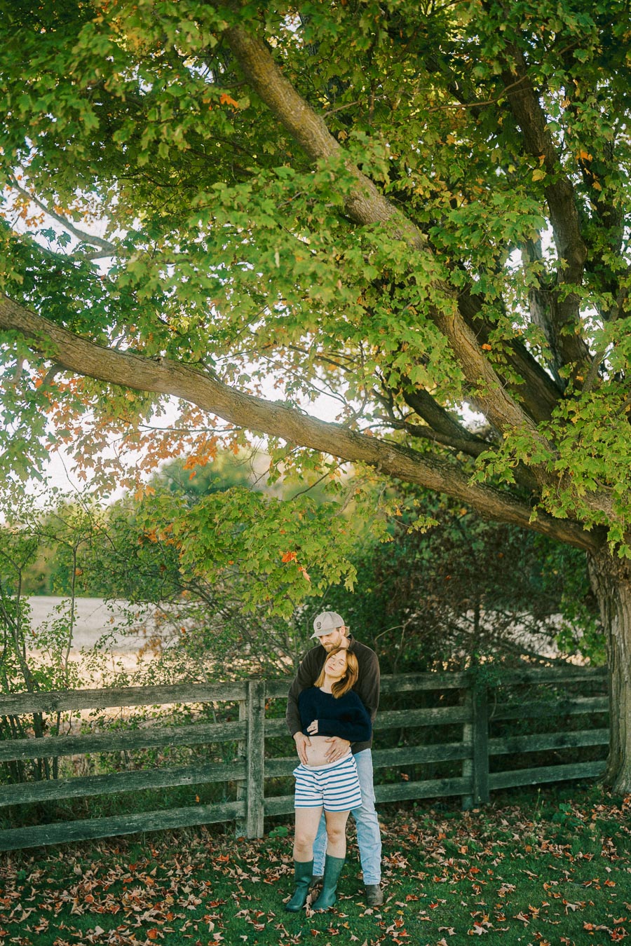 A couple embracing under a large, leafy tree in a park, with a rustic wooden fence in the background. The ground is covered with fallen autumn leaves, creating a serene and romantic atmosphere.