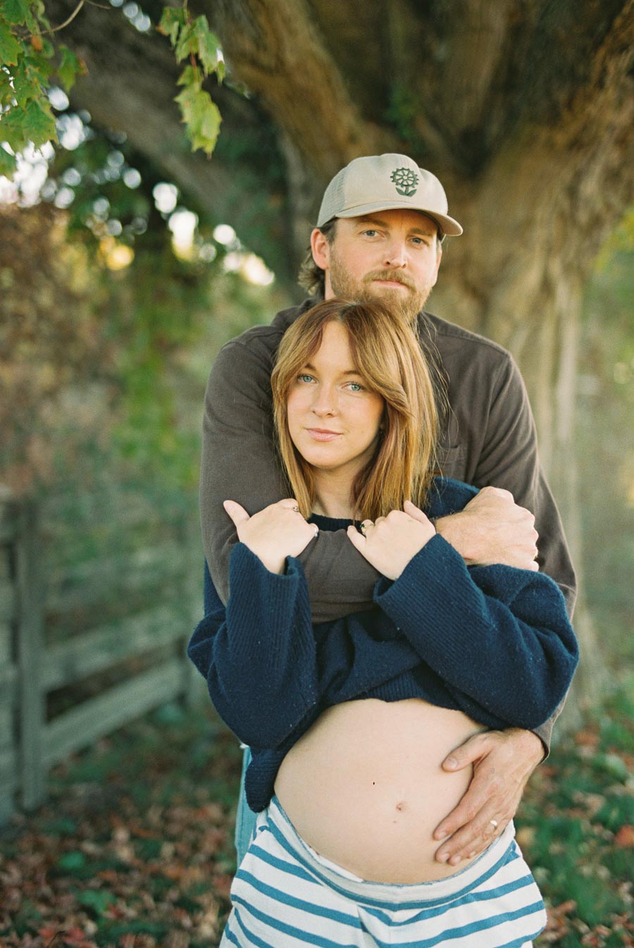A couple embracing outdoors with a focus on the woman's pregnant belly, surrounded by lush greenery and under a large tree.