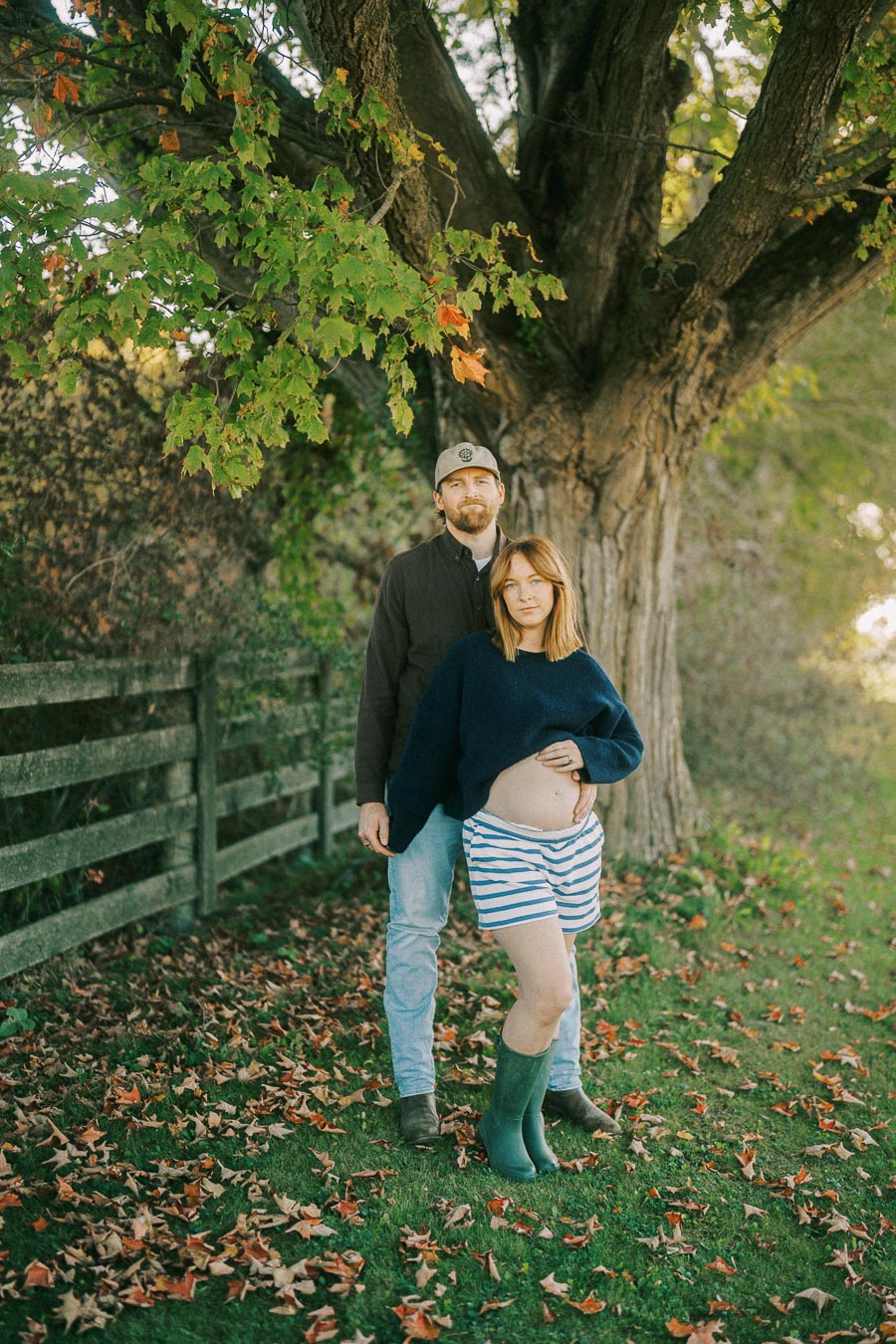 Pregnant woman in striped shorts and green boots, standing with a partner under a large tree with autumn leaves on the ground.