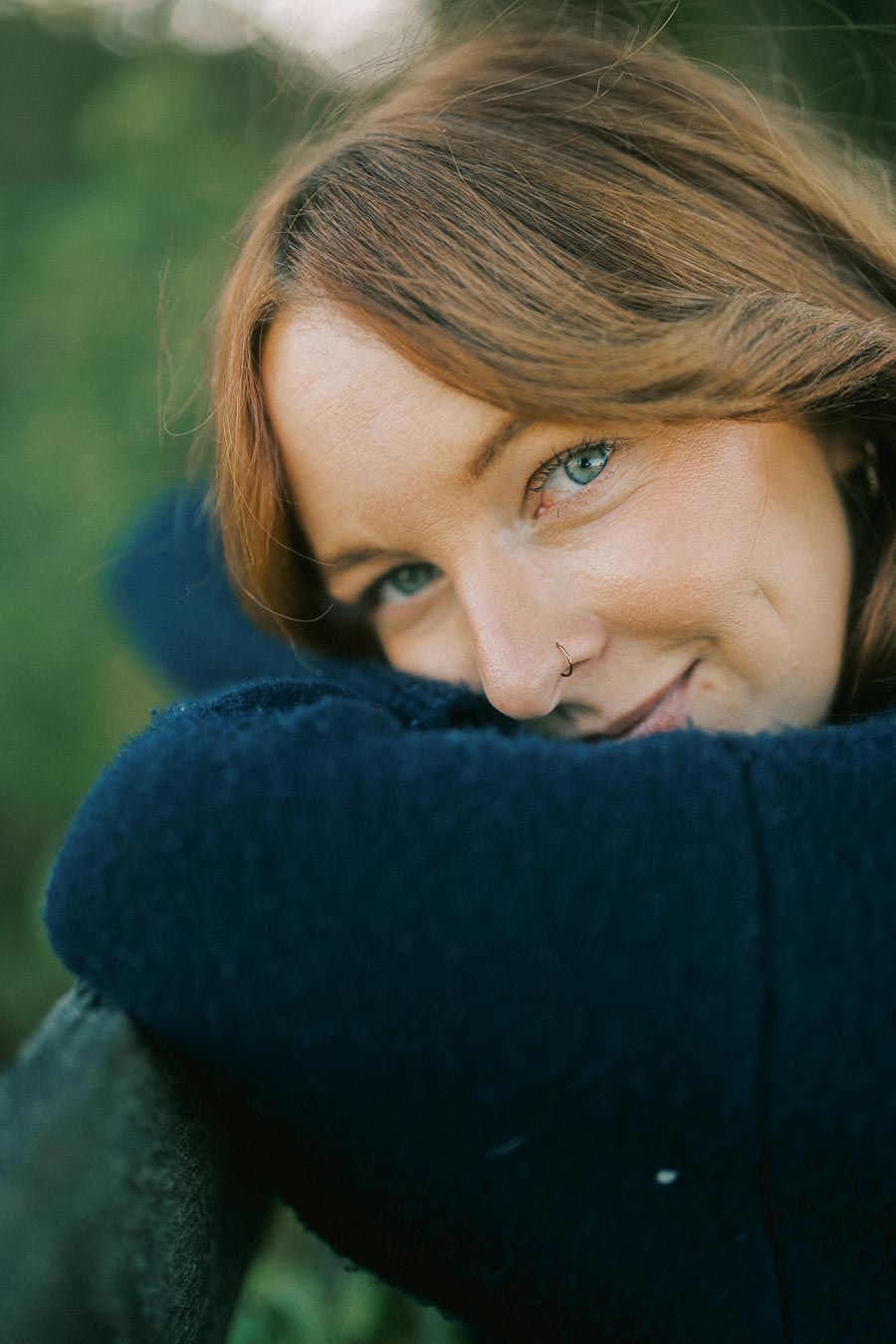 Close-up of a person with blue eyes and auburn hair, wearing a dark blue sweater, leaning on an outdoor railing, surrounded by greenery.