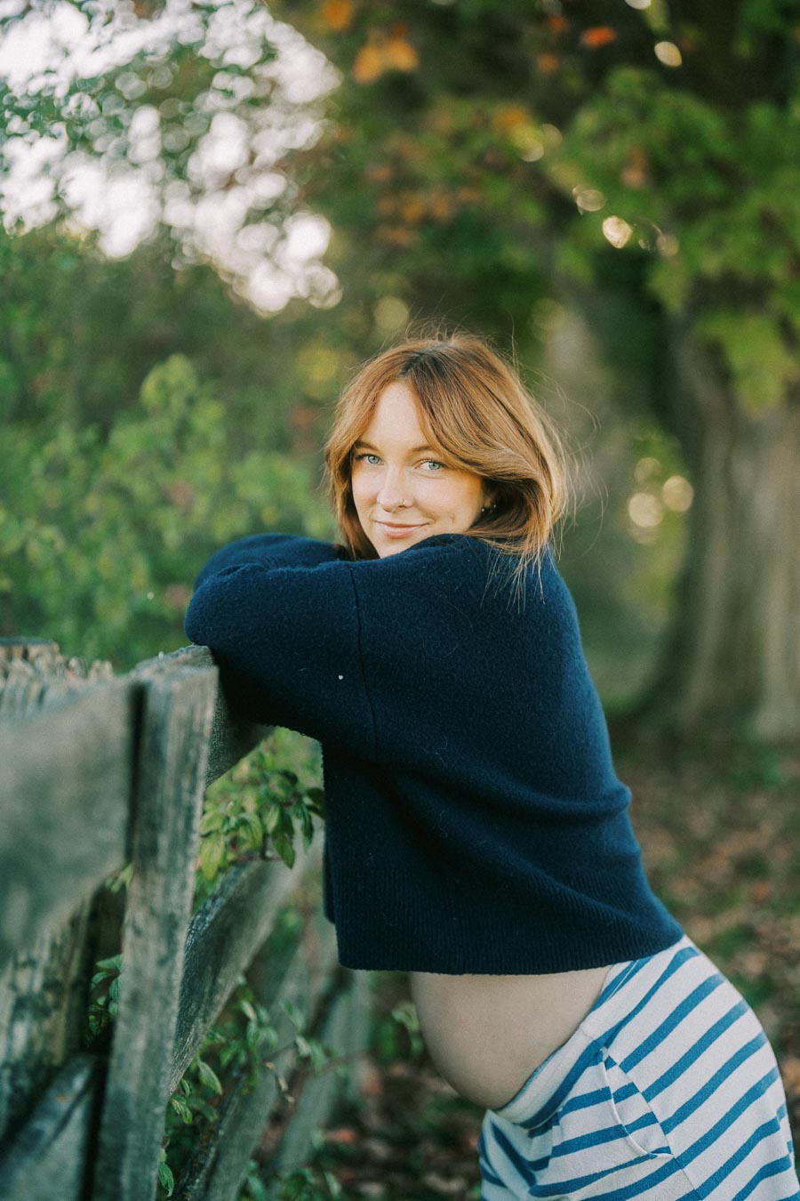 A pregnant woman in a navy sweater and striped pants leaning against a wooden fence, surrounded by lush greenery and autumn foliage, with a serene expression.