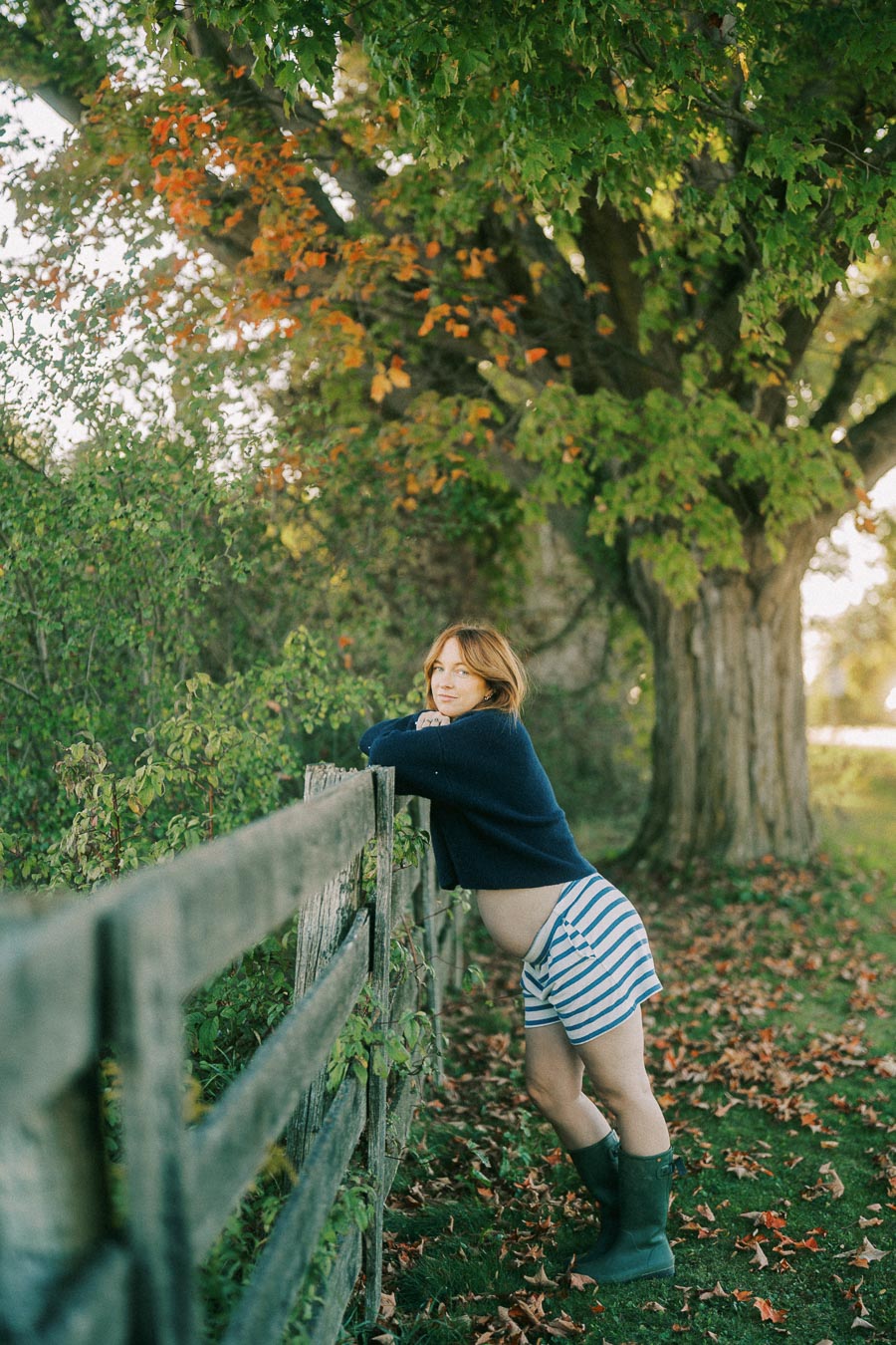Pregnant woman leaning on a wooden fence in a grassy field, surrounded by autumn leaves and trees, wearing a blue sweater and striped shorts.