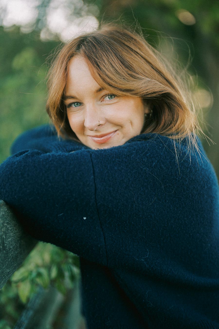 Smiling woman with brown hair leaning on a fence in a cozy blue sweater, surrounded by greenery