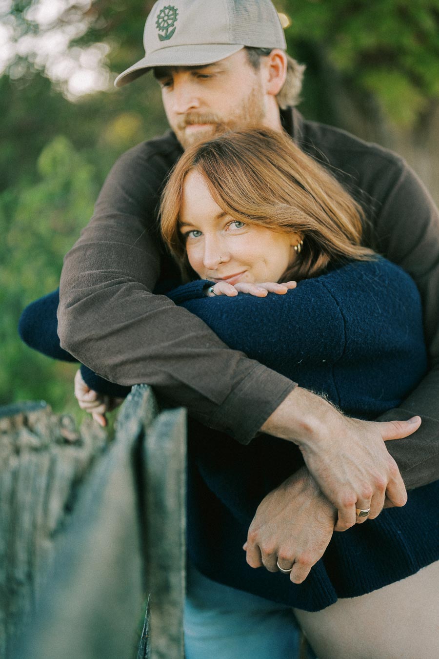 A couple embracing outdoors with a wooden fence in the foreground, surrounded by greenery. The woman smiles while the man in a cap gently holds her from behind.