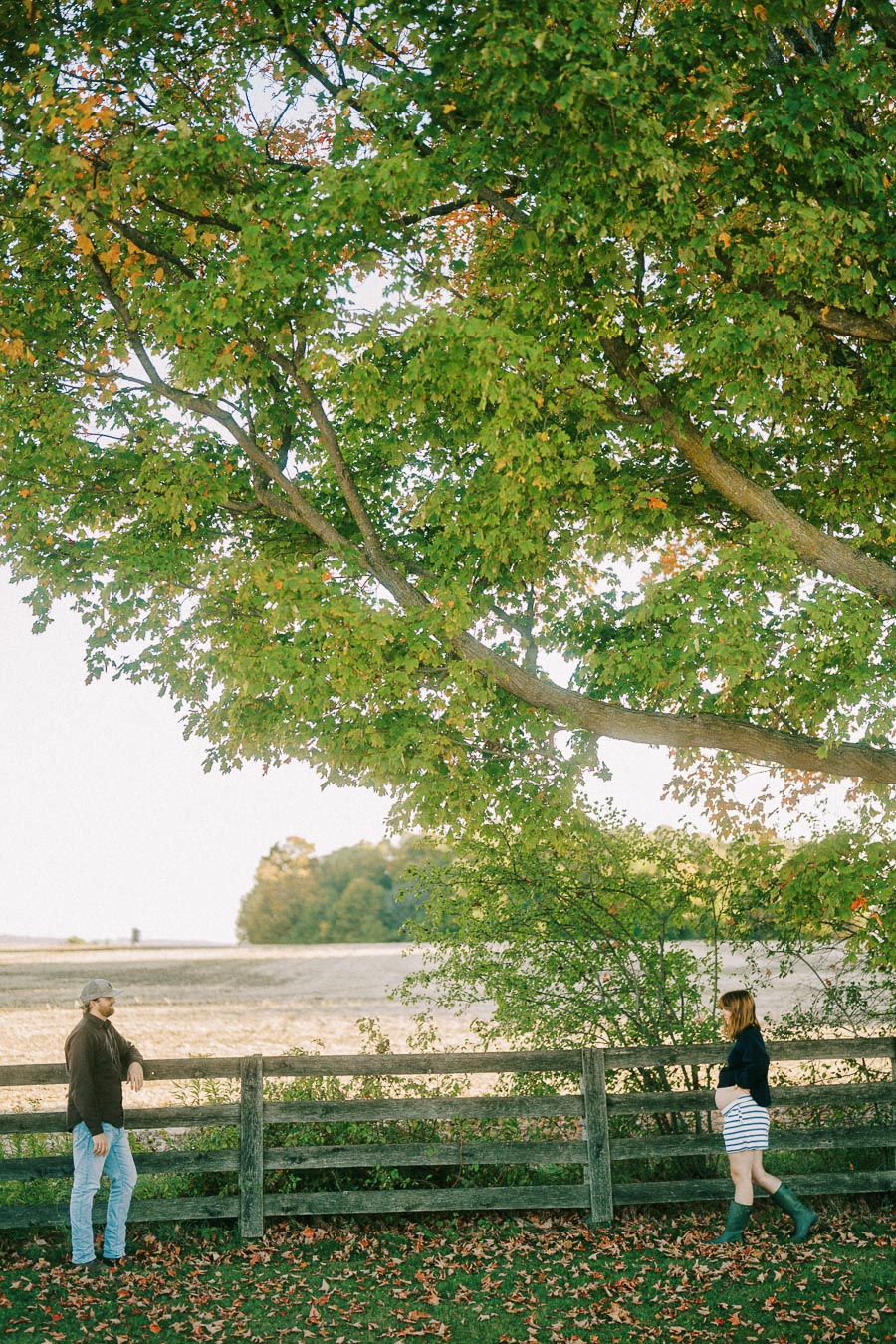 A man and a woman enjoy a scenic autumn walk by a wooden fence under a large tree, with fallen leaves covering the ground and a clear sky in the background.