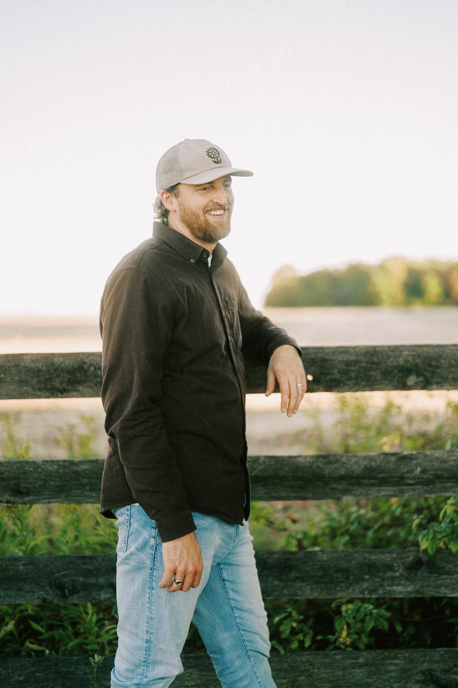 Man wearing a brown jacket and cap leaning on a wooden fence in a countryside setting, smiling against a clear sky background.