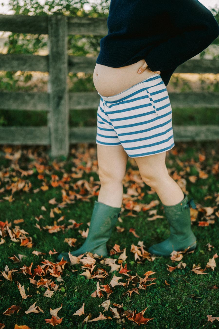 Pregnant person in striped shorts and green boots standing outdoors among fallen autumn leaves.