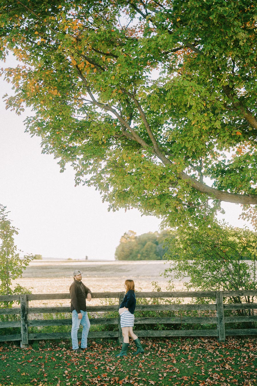 A couple standing by a rustic wooden fence under a large tree with green and orange leaves, on an autumn day in the countryside.