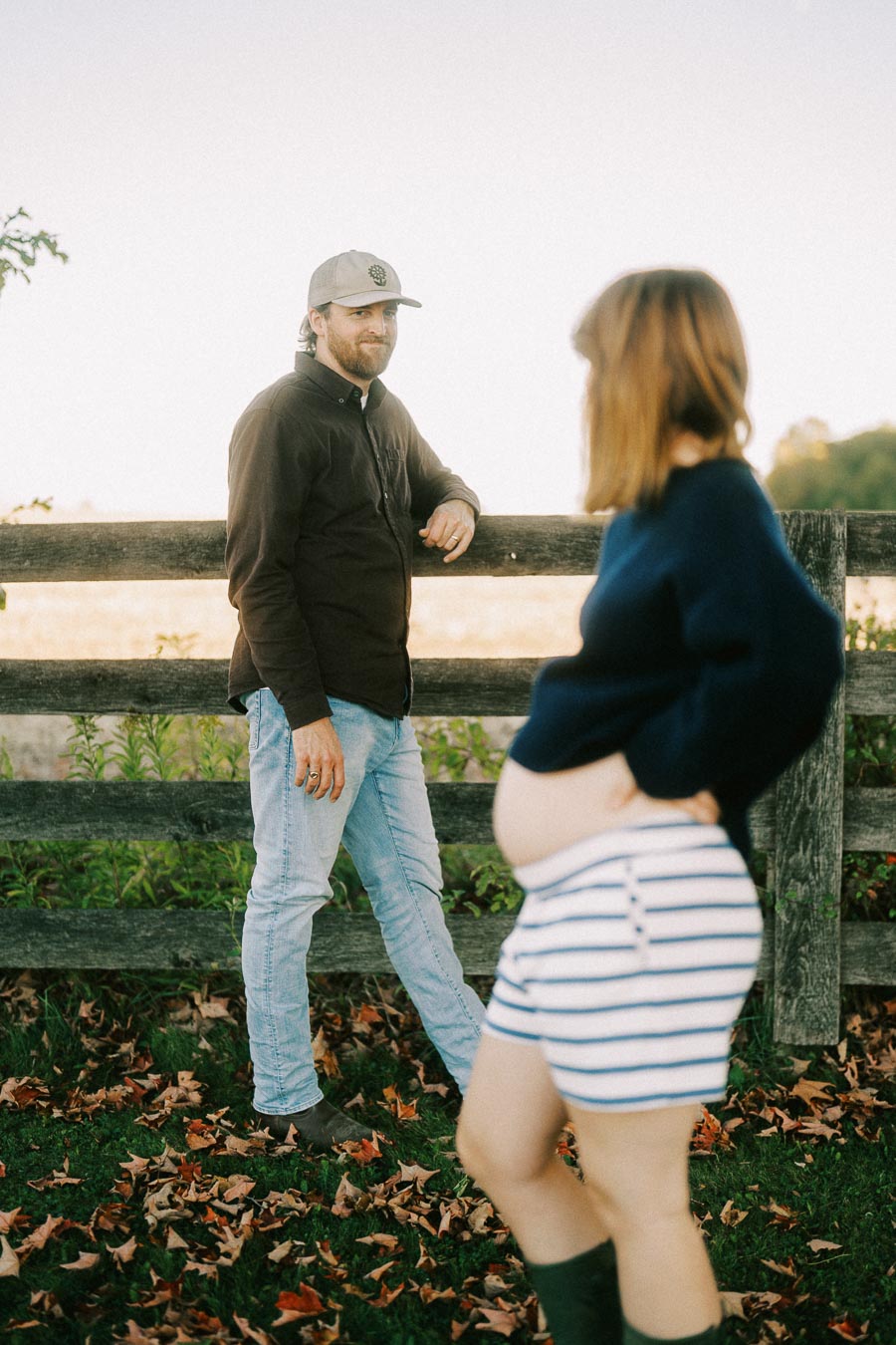 Man in brown jacket and jeans leaning on a wooden fence, looking at a pregnant woman in a navy sweater and striped shorts standing on grass covered with autumn leaves.