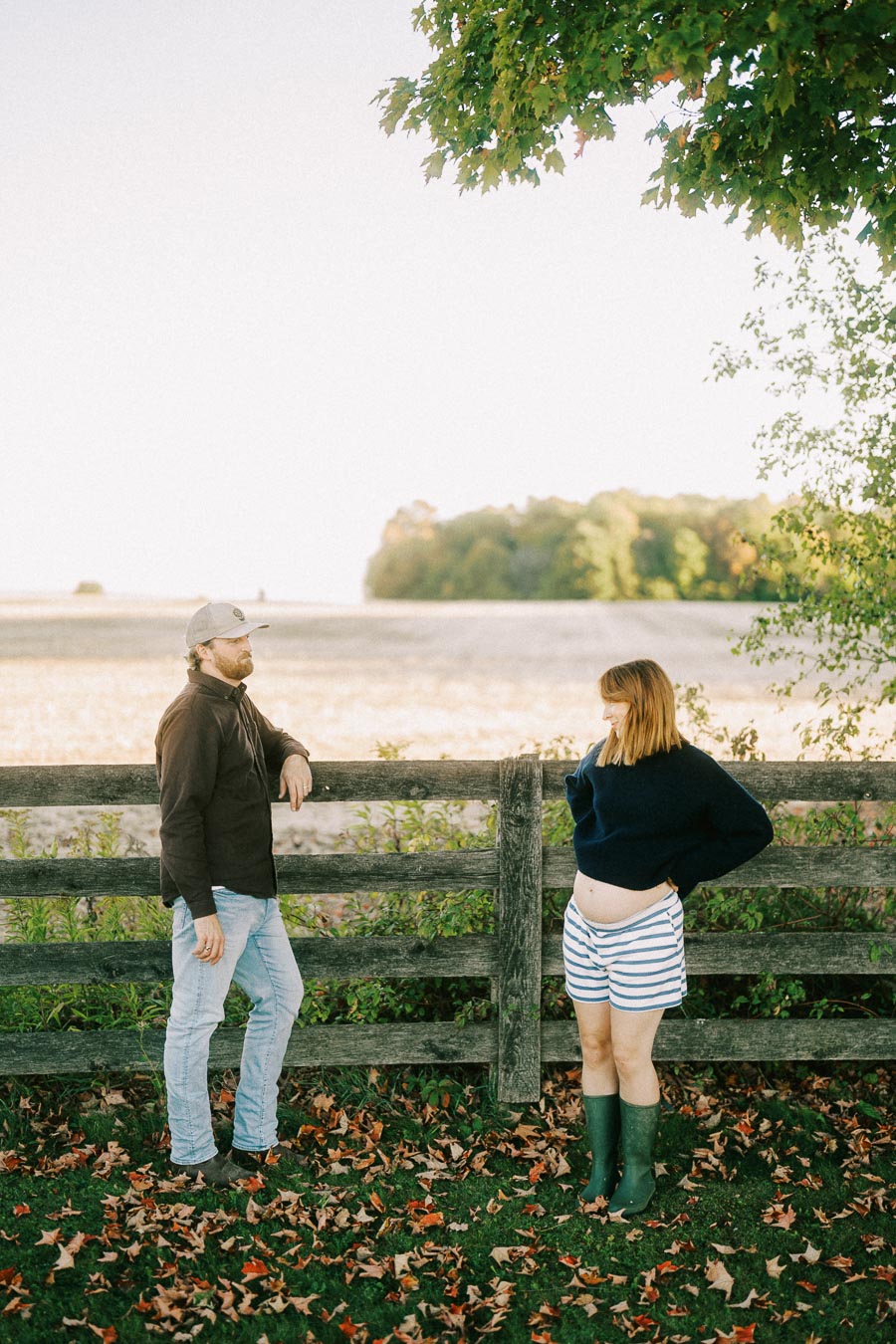 A man wearing a cap and jeans leans on a wooden fence while conversing with a pregnant woman in a striped outfit and boots on a leafy autumn day in a rural setting.
