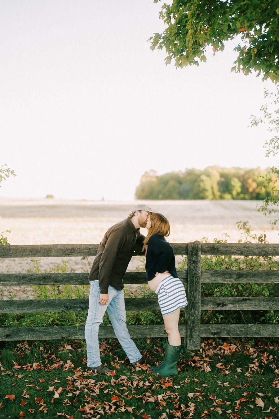 A couple kissing by a wooden fence with autumn leaves on the ground, featuring a pregnant woman in a striped skirt and green boots, under a canopy of trees.