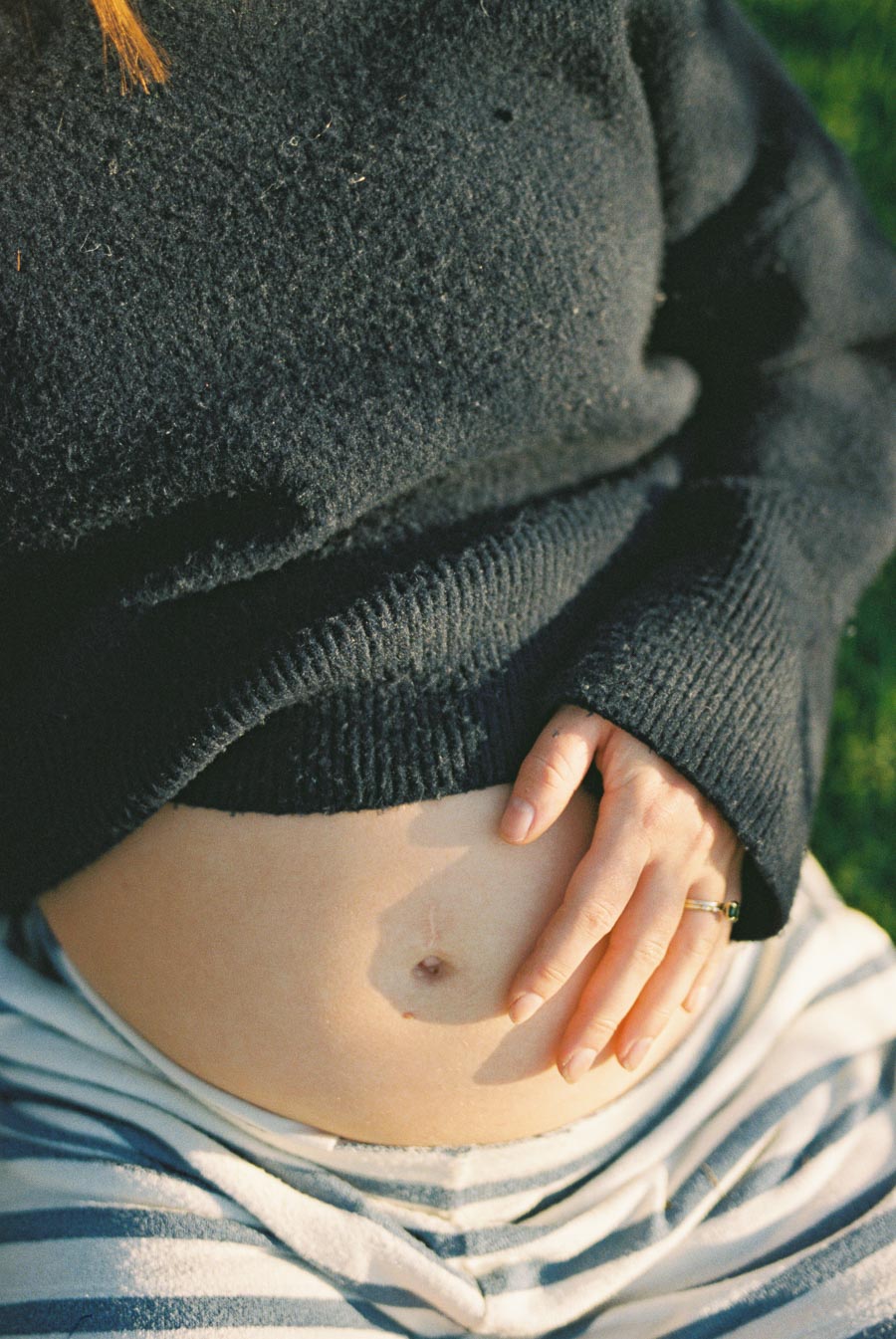Pregnant woman resting hand on bare belly, wearing a dark sweater and striped pants, sitting outdoors on a sunny day.