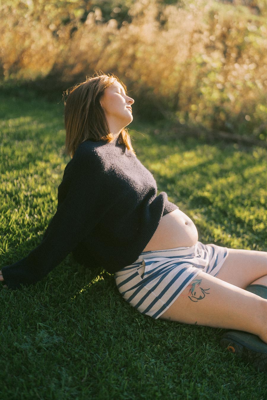 A pregnant woman relaxing on grass, wearing a dark sweater and striped shorts, enjoying sunlight in a serene outdoor setting.