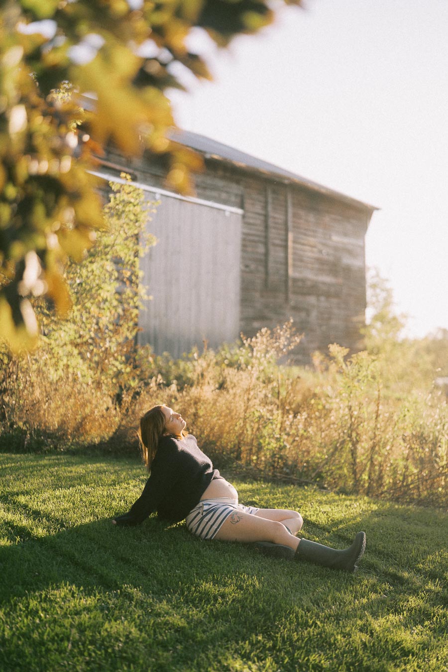 Pregnant woman relaxing on grass near rustic wooden barn under sunlight.