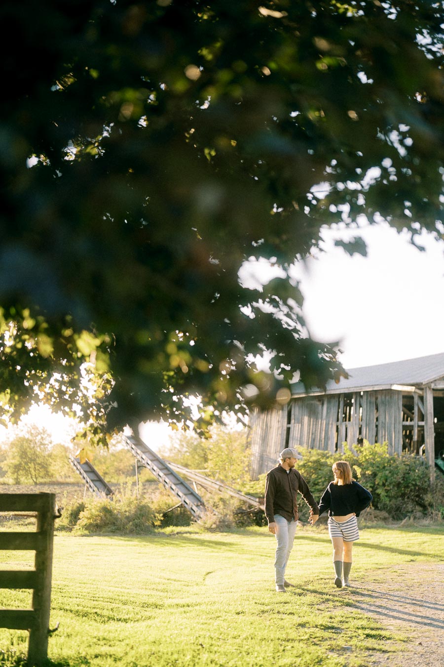 A couple walking hand in hand through a sunny rural landscape with an old barn in the background, surrounded by lush green grass and trees.