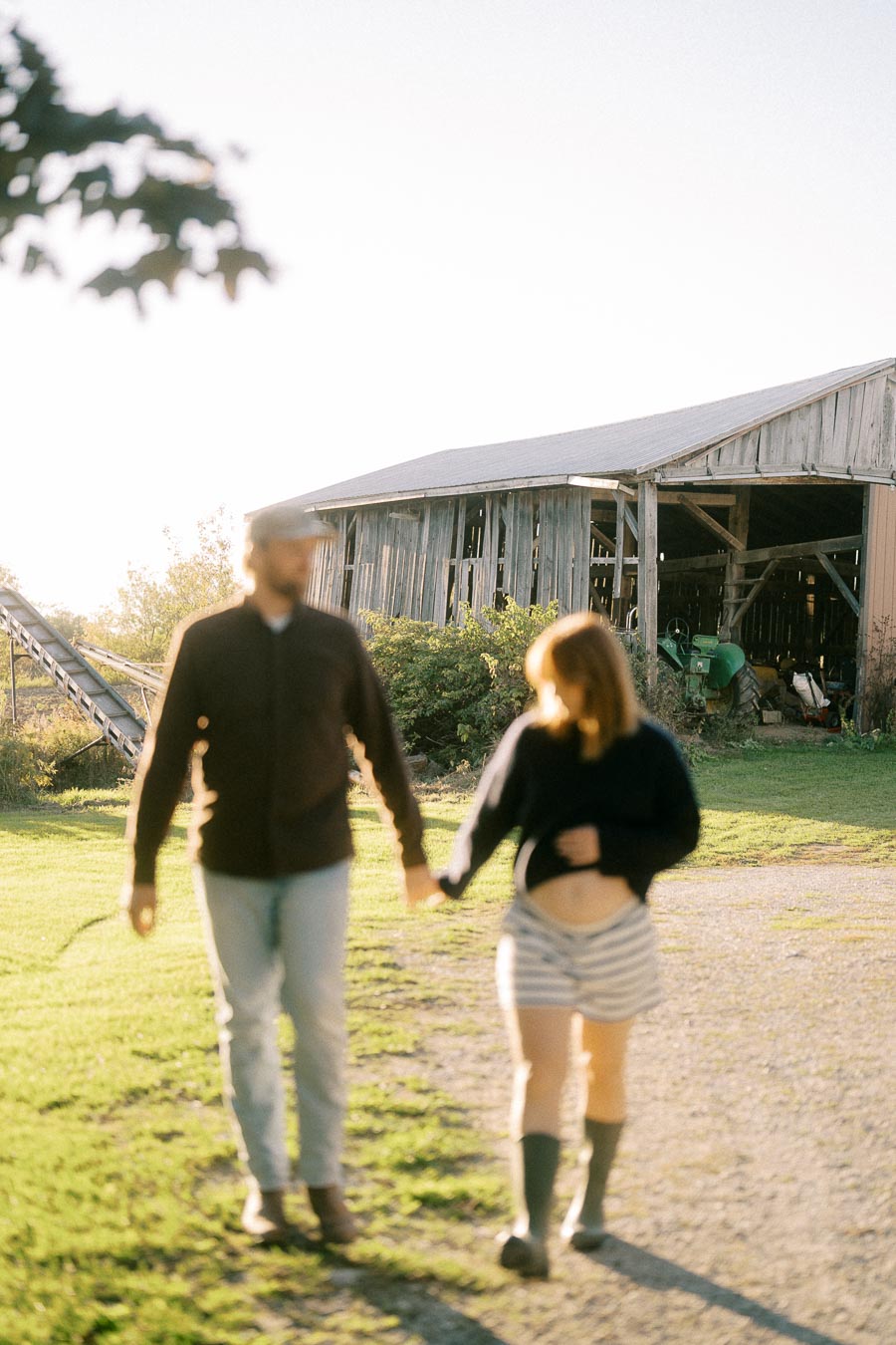 A couple holding hands walks on a sunny day near a rustic barn and green tractor, set against a rural landscape.