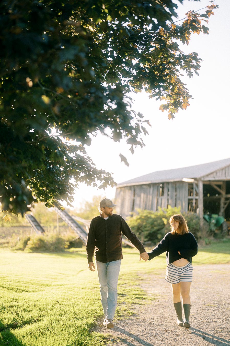 Couple walking hand in hand on a sunlit path near a rustic barn, surrounded by lush greenery.