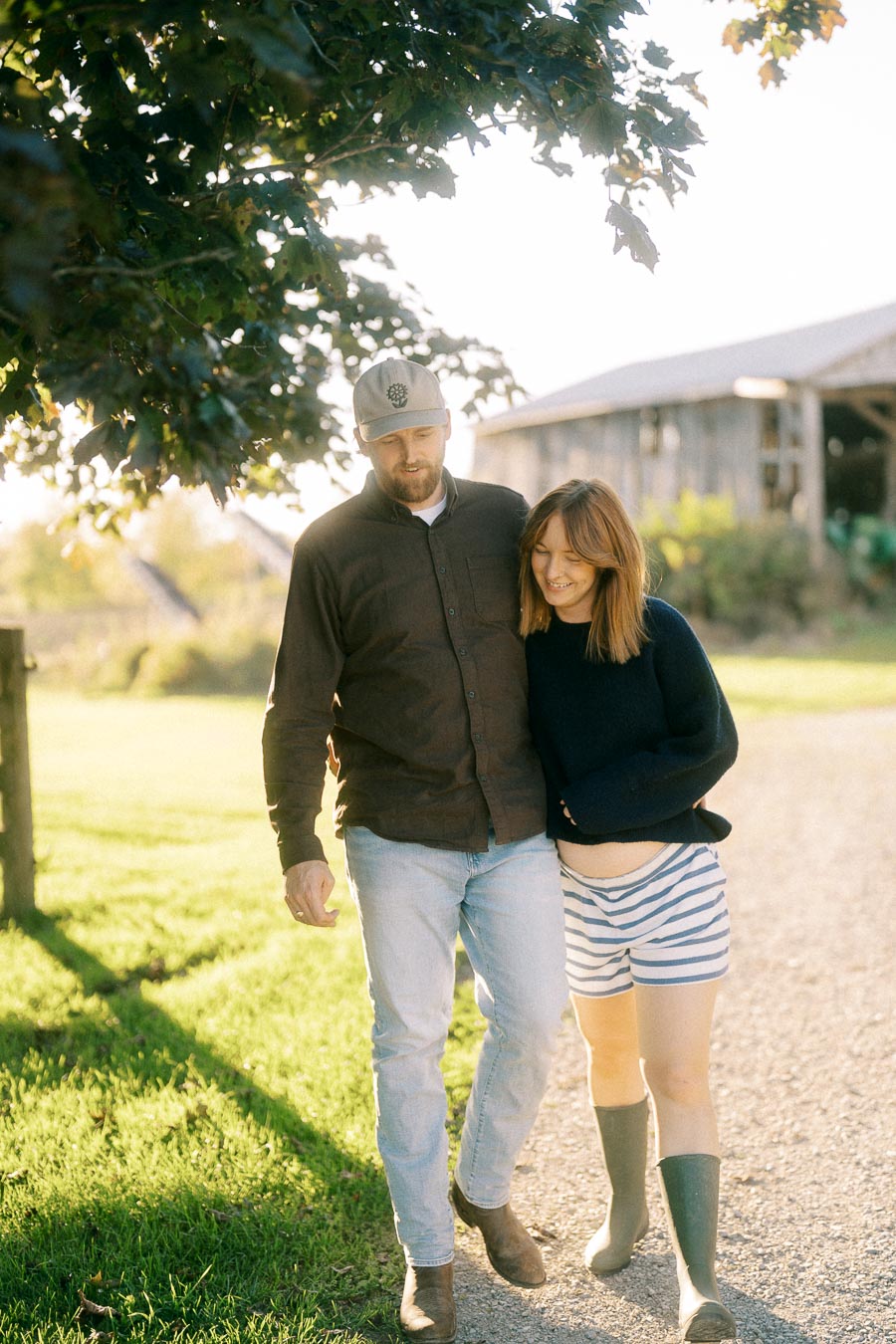 A couple walking on a sunny farm path, surrounded by greenery, wearing casual clothes and rubber boots, with a rustic barn in the background.