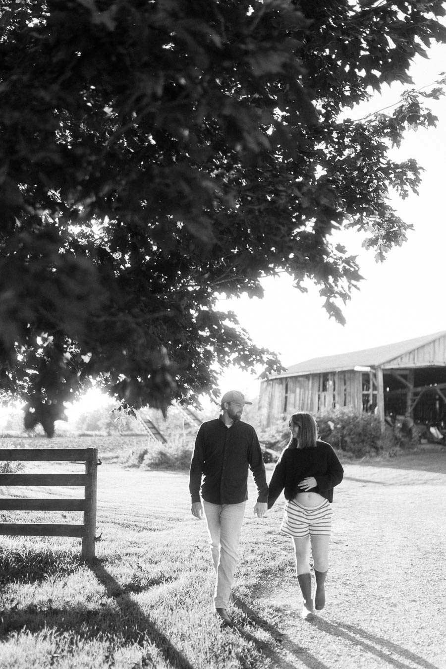 A couple holding hands and walking under a large tree in a rural setting with a rustic barn in the background, captured in black and white.