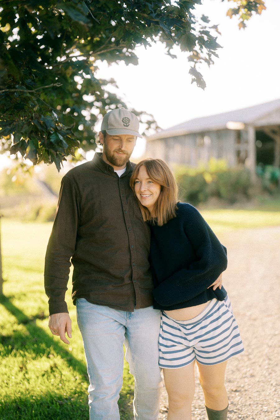 A happy couple enjoying a sunny day outdoors, standing close together under a tree with a rustic barn in the background. The man is wearing a cap and a brown shirt, while the woman is in a navy sweater and striped shorts, smiling warmly.
