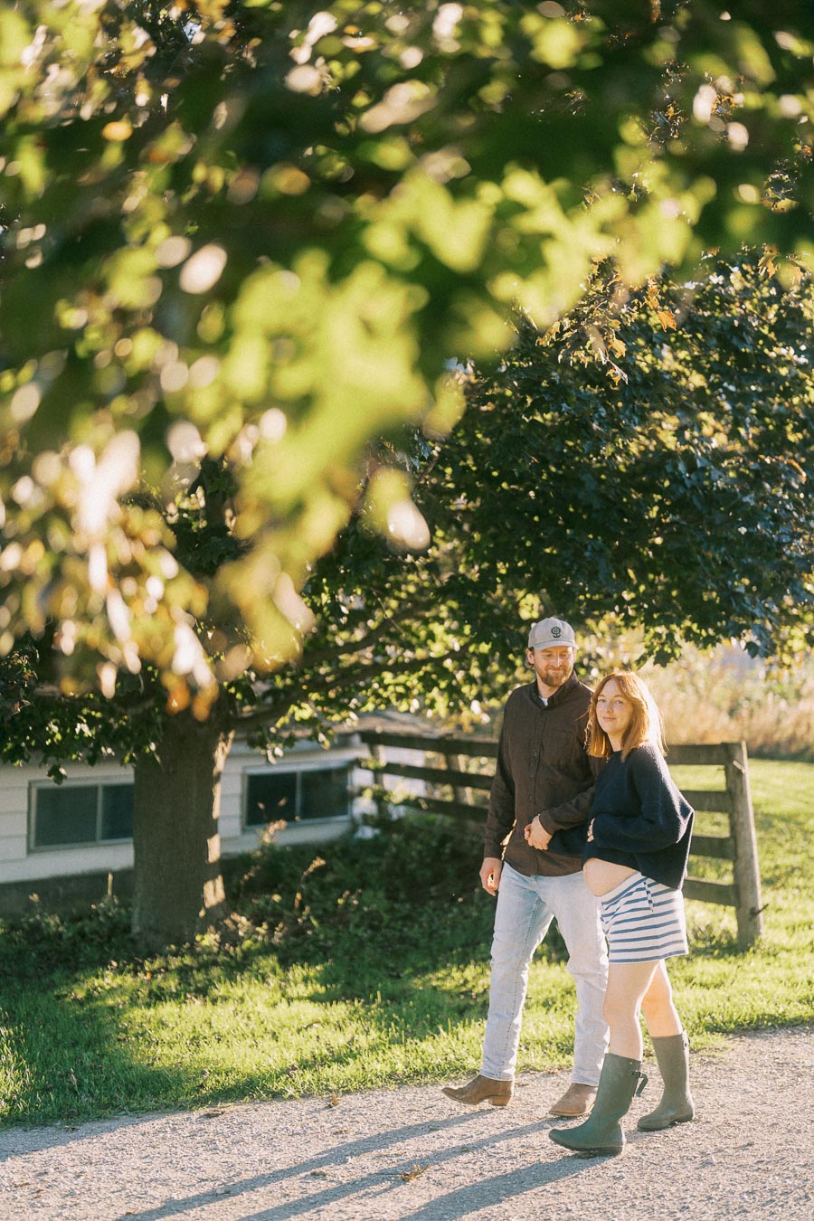 A couple walking arm in arm on a sunny day, with a pregnant woman smiling and touching her belly, wearing casual outdoor clothing near a wooden fence and lush greenery.