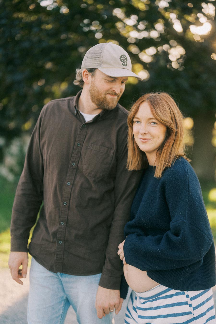 A couple enjoying a sunny day outdoors, with the woman showing off her pregnancy in a stylish blue sweater and striped pants. The man stands beside her affectionately, wearing a cap and casual outfit, as sunlight filters through the trees in the background.