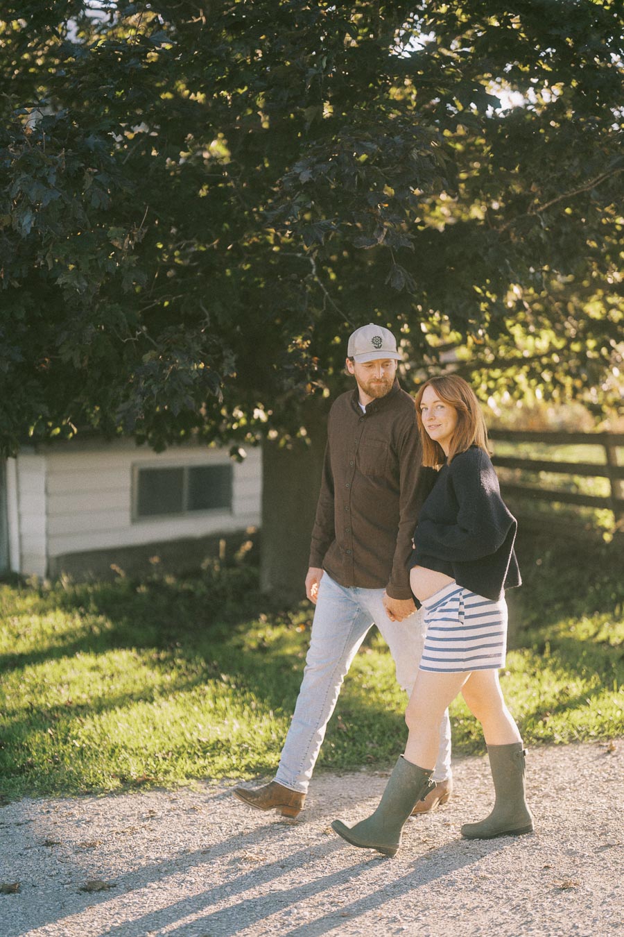 A couple walking hand in hand outdoors, with the woman visibly pregnant, wearing a striped dress and boots, surrounded by lush greenery and sunlight filtering through the trees.