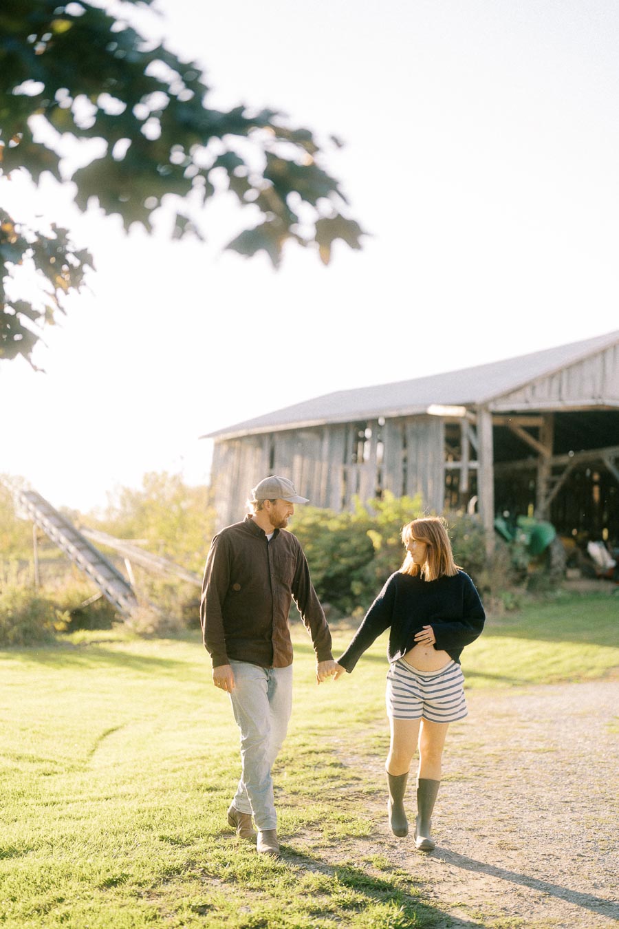 A couple walking hand in hand on a sunny farm, wearing casual clothing and boots, with a rustic barn in the background.