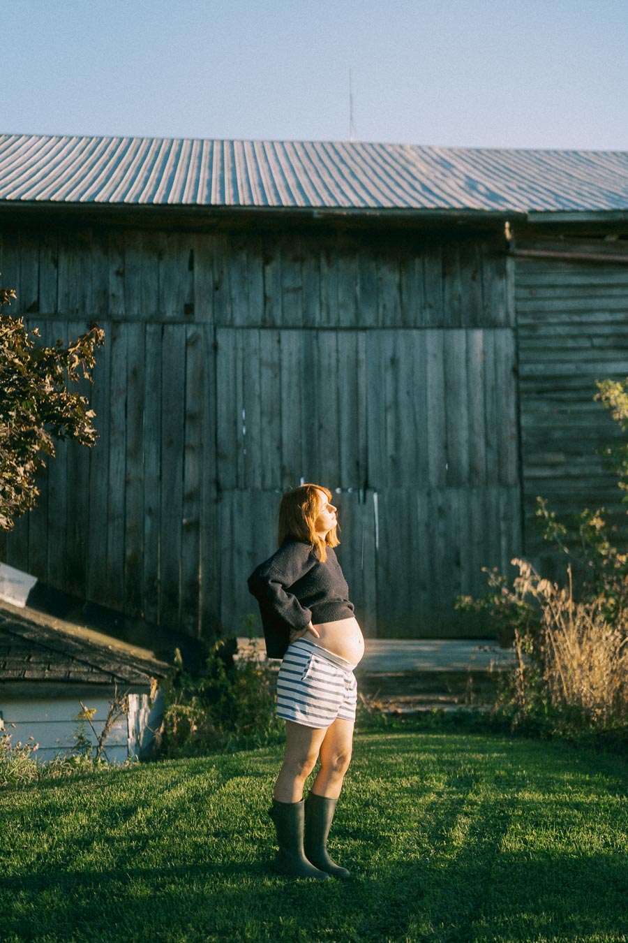Pregnant woman in striped shorts and boots standing on grass, in front of a rustic wooden barn, enjoying sunshine.
