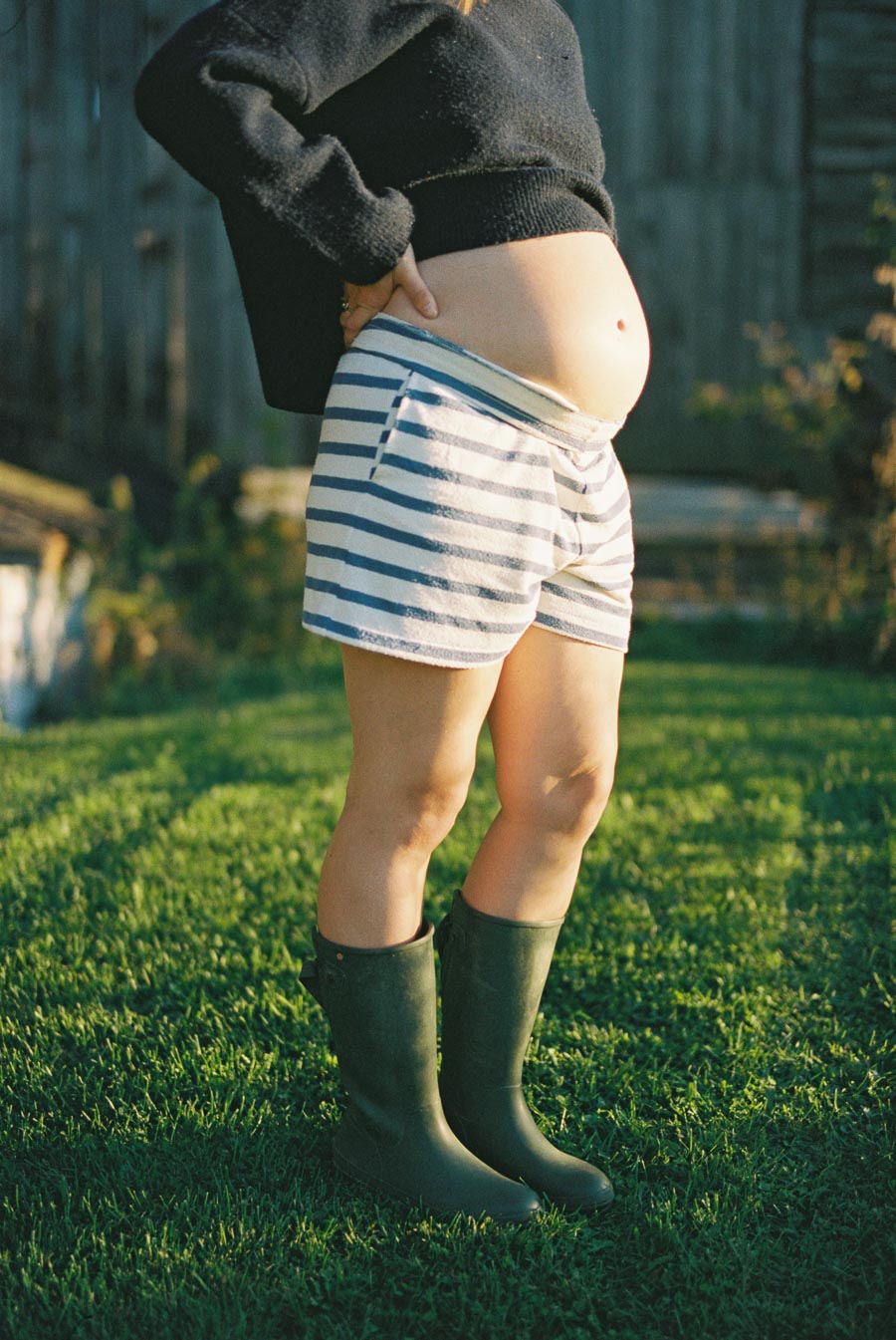 Pregnant woman in cozy sweater and striped shorts standing on grass, wearing rubber boots, in a sunny garden setting.