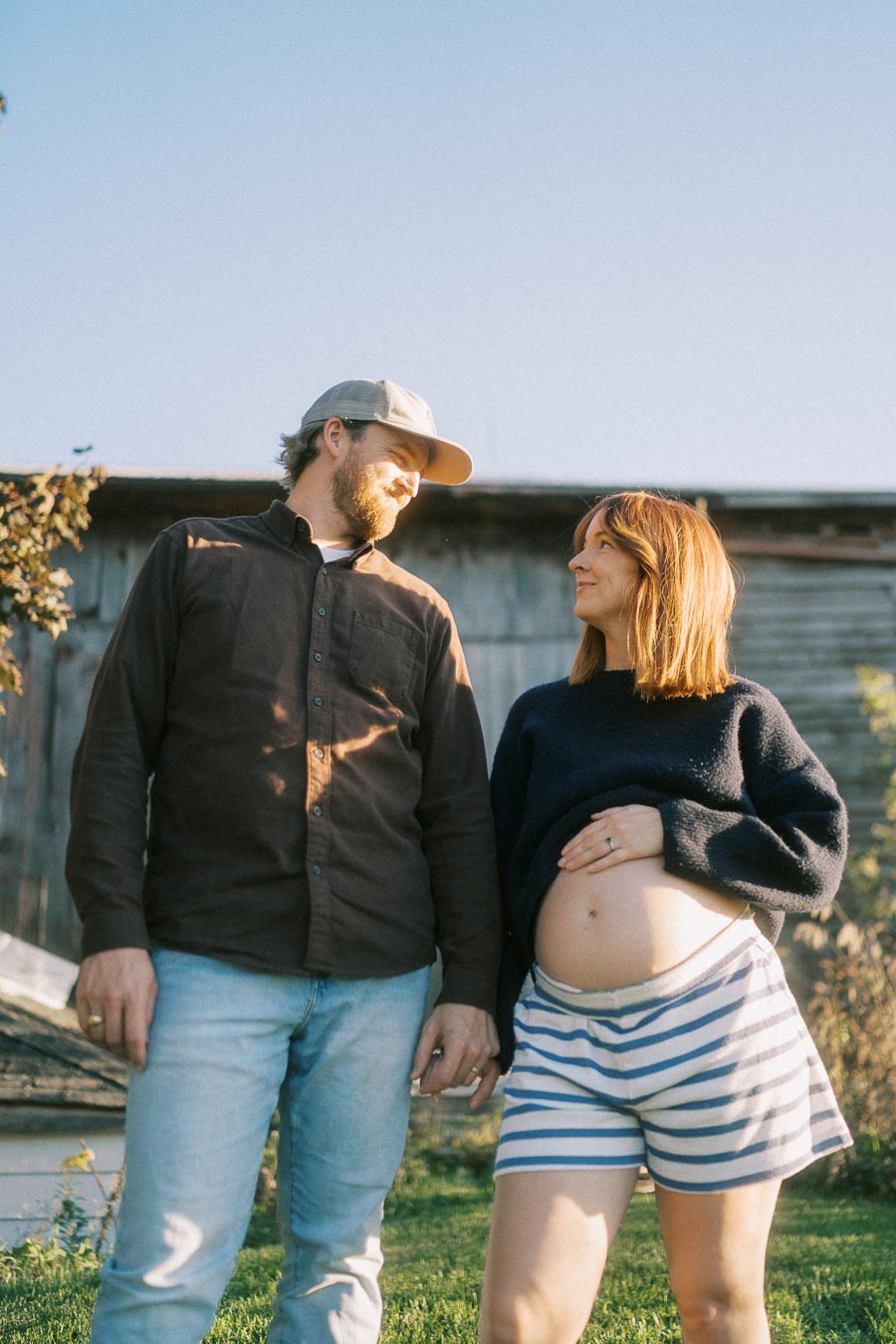 Happy couple standing in a sunny yard, with the woman showcasing a pregnant belly, wearing casual clothes and holding hands, with a rustic barn in the background.
