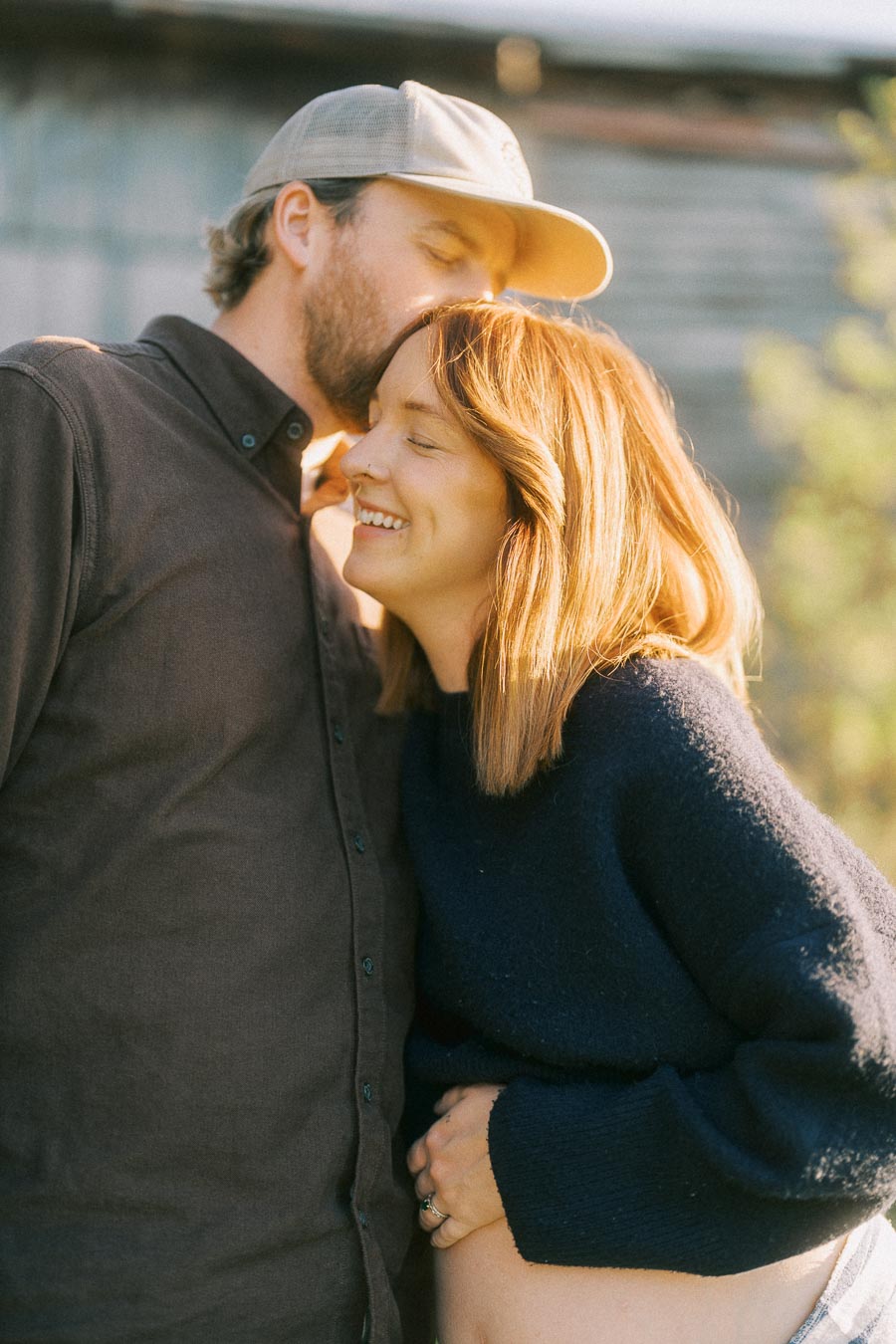 Couple sharing a tender moment outdoors, with man in cap kissing woman's head as she smiles, highlighting joy and intimacy.